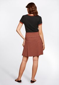 Woman standing with back turned wearing a black short-sleeve shirt and a red patterned knee-length skirt with black sandals.