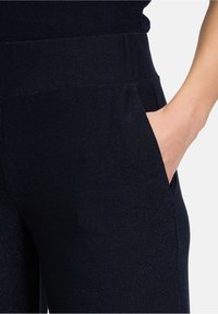 Marineblaue Hose mit weicher Textur, breitem Bund und seitlichen Taschen. Dezente metallische Fäden sind im gesamten Stoff sichtbar.