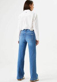 Eine Frau steht mit dem Rücken zur Kamera, trägt ein weißes Langarmshirt, blaue gerade Jeans und beige Schuhe vor einem schlichten weißen Hintergrund.