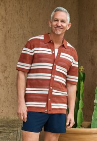 Short-sleeve button-up shirt in rust with white stripes, featuring a textured knit design. Paired with navy shorts, displayed outdoors.
