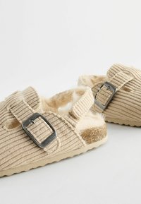 Beige kordflorsandaler med fuskpälsfoder, justerbar rem och silverfärgad metallspänne. Texturerad ovandel och robust, flexibel sula.