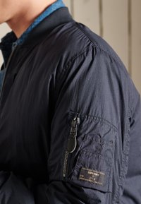 Close-up of a person wearing a navy blue Superdry jacket with a zippered pocket and label on the upper arm.