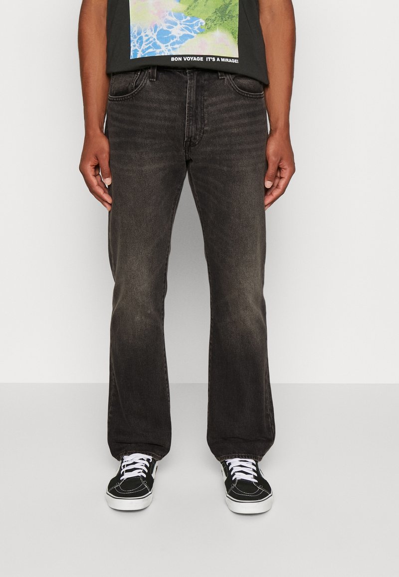 Homme portant un jean noir délavé à jambes droites et des baskets noires à lacets blancs, debout devant un fond clair uni.