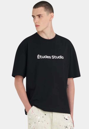 Jeune homme aux cheveux courts portant un T-shirt noir ample avec le texte "Études Studio" et un pantalon clair éclaboussé de peinture, les mains dans les poches.