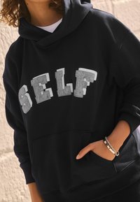 Schwarzer Kapuzenpullover mit einem großen "SELF"-Druck in Grau und Weiß, ausgestattet mit einer Kängurutasche und gerippten Bündchen. Weicher Stoff und lockere Passform.