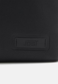 Bolso de cuero negro con una superficie texturizada y un prominente parche rectangular de logo con el nombre de la marca "Jost."