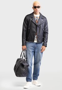 Veste en cuir noir de motard avec zipper asymétrique, pull à motifs en dessous, jean bleu et sac de sport en cuir noir. Baskets blanches.