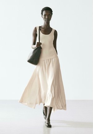 Donna che cammina in avanti indossando una maglietta senza maniche beige, gonna fluente color crema, scarpe basse nere e portando una borsa nera a tracolla su uno sfondo chiaro.