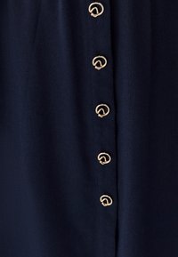 Tessuto blu navy dalla texture liscia con bottoni dorati a forma circolare lungo la cucitura verticale, che aggiungono un dettaglio decorativo al capo.