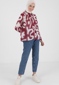 Rot und weiß gemusterte Bluse mit hohem Kragen, Knopfverschluss und lockeren Ärmeln, kombiniert mit blauen Jeans und weißen Turnschuhen.