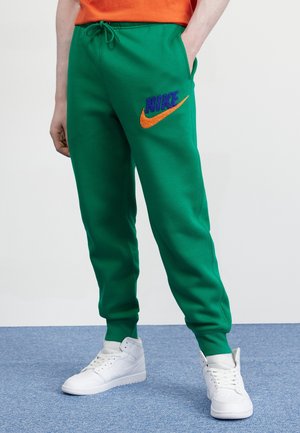 Personne portant un pantalon de survêtement vert Nike avec un logo orange et bleu et des baskets montantes blanches, debout sur un tapis bleu.