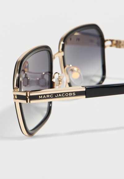 Γυαλιά ηλίου με τετράγωνα πλαίσια σε χρυσό και μαύρο χρώμα, με βαθμωτούς σκούρους φακούς και "MARC JACOBS" τυπωμένο στον βραχίονα του πλαισίου.