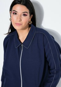 Giovane donna con capelli scuri che indossa orecchini a cerchio dorati e una giacca blu navy con cerniera e cuciture bianche, su sfondo bianco.