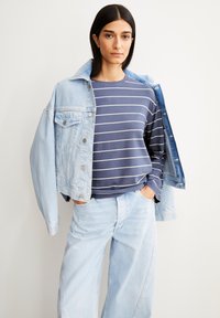 Denimjacke mit heller Waschung, offen über einem navy-blauen, gestreiften Langarmshirt getragen. Helle, weit geschnittene Jeans vervollständigen das Outfit.
