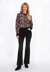 Donna in piedi con una mano in tasca, indossa un maglione paisley nero e rosa, jeans neri a zampa d'elefante e stivali neri con tacco.