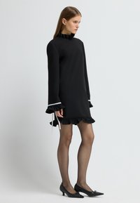 Černé šaty s dlouhými rukávy a vysokým volánkovým límcem, volánkovým lemem a manžetami, s bílými akcenty. V kombinaci s černými lodičkami se špičatou špičkou.