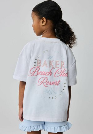 Mädchen mit dunklem, lockigem Haar im Pferdeschwanz trägt ein weißes, übergroßes T-Shirt mit dem Schriftzug "Baker Beach Club Resort" und hellblaue Rüschen-Shorts.