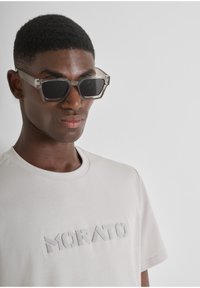 Gafas de sol transparentes con forma hexagonal y montura transparente, llevadas sobre una camiseta gris claro con letras bordadas texturizadas que dicen "MORATO".