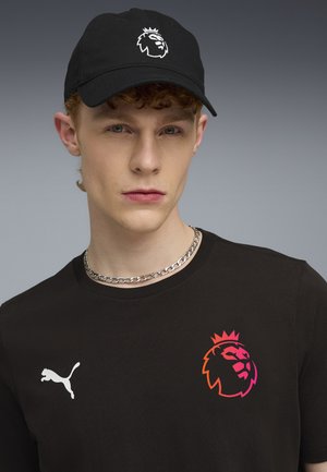 Czarna czapka z logo lwa, czarna koszulka z krótkim rękawem z emblematem lwa w gradientowym kolorze oraz srebrny naszyjnik z łańcuszkiem.