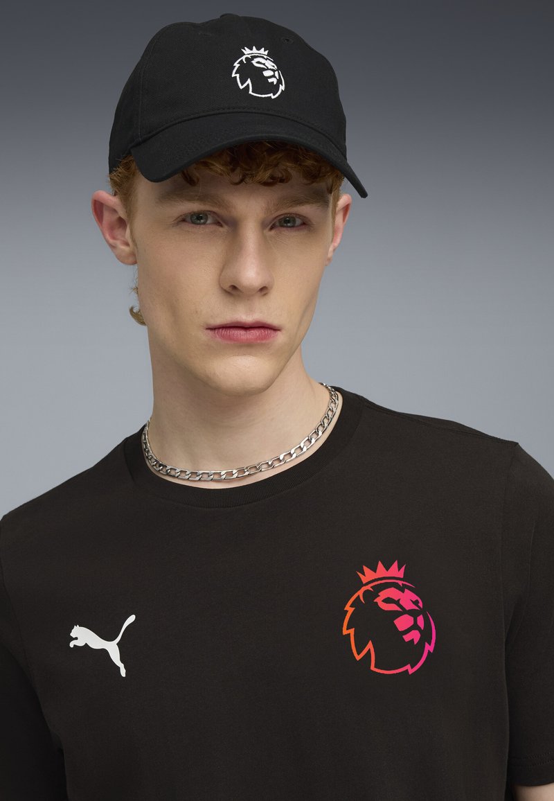 Casquette noire avec logo de lion, chemise noire à manches courtes arborant un emblème de lion en dégradé, et collier chaîne argenté.