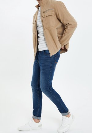 Homme portant une veste beige sur une chemise rayée, un jean bleu retroussé et des baskets blanches, debout avec une main dans la poche sur fond blanc.