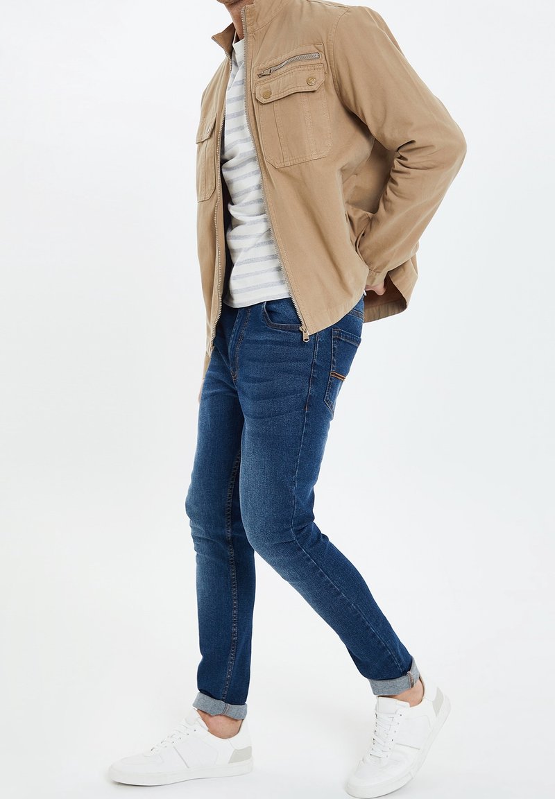 Homme portant une veste beige sur une chemise rayée, un jean bleu retroussé et des baskets blanches, debout avec une main dans la poche sur fond blanc.