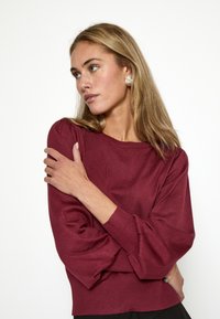 Burgunderfarbener Strickpullover mit geripptem Saum, gepufften Ärmeln und weitem Halsausschnitt. Glatte Textur und figurbetontes Design.