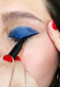 Un eye-liner noir appliqué sur une paupière ornée d'ombreuse bleue vive, avec un gros plan sur des ongles peints en rouge.