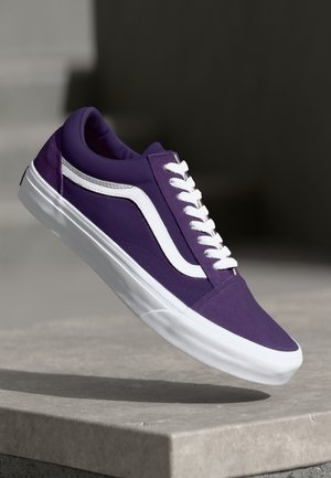 Baskets violettes avec une bande blanche et des lacets flottant au-dessus d'une surface en pierre sur un fond sombre et neutre.