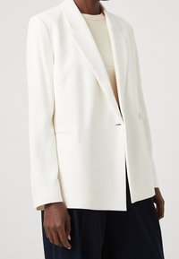 Blazer blanco a medida con un solo botón, solapa de muesca y una textura suave, llevado sobre una blusa beige clara y pantalones azul marino.