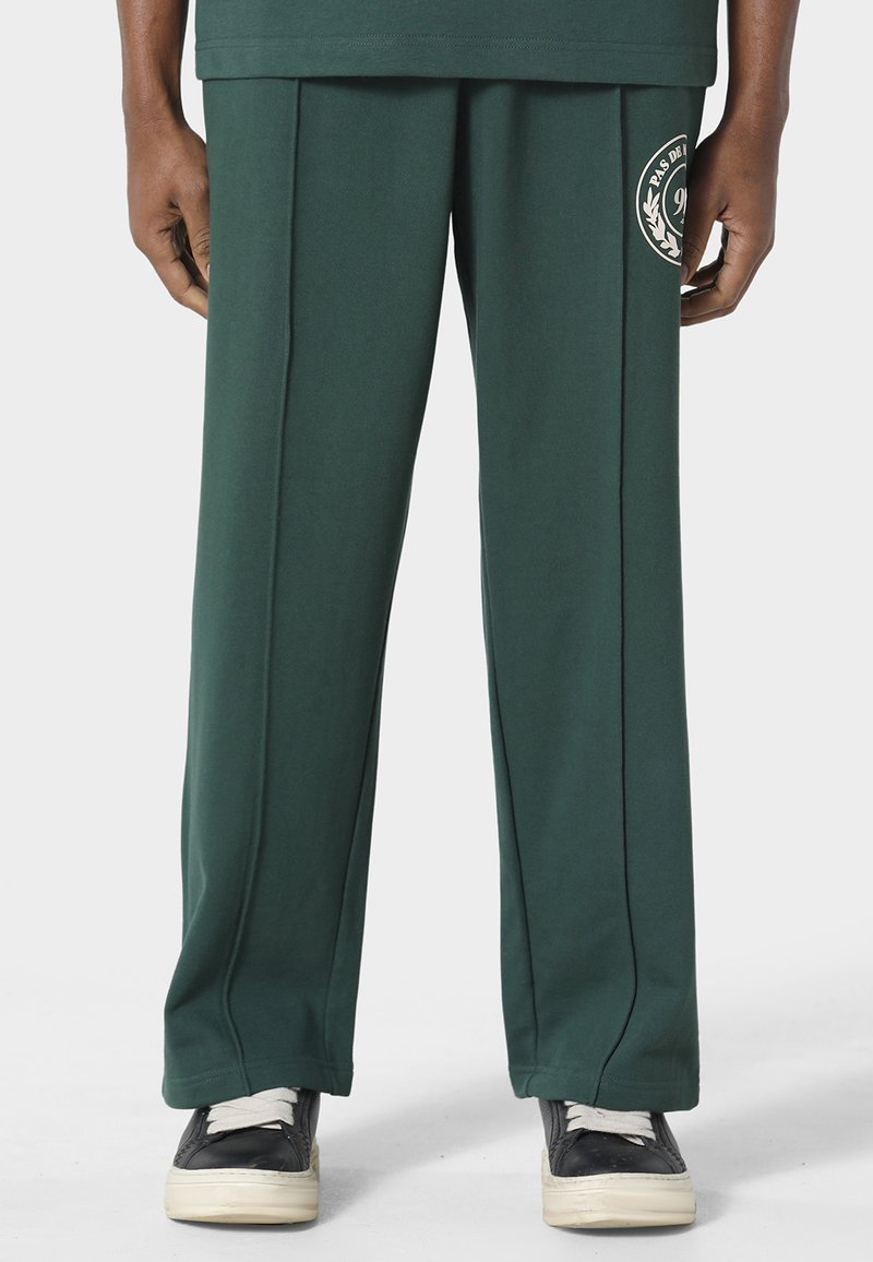 Personne portant un pantalon ample vert foncé avec des détails de couture à l'avant et des baskets noires à lacets blancs sur un fond uni.