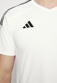 Camiseta deportiva blanca con rayas negras en los hombros y un logotipo bordado en negro en la parte superior izquierda del pecho. Textura de tela suave.