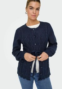 Mornarica pletena kardigan s teksturiranim dijamantskim uzorkom, rebrastim rukavima, pet gumba i vezicom u struku. Detalji s valovitim rubom.