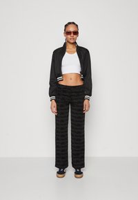 Zwarte nylon bomberjack met witte strepen, witte crop top, zwarte gestreepte wijde broek en zwarte sneakers met lichte zolen.