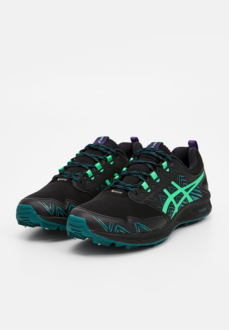 Zapatillas de running negras con acentos verdes, parte superior de malla texturizada, patrones geométricos y suela resistente. Incluye cordones y detalles de refuerzo.