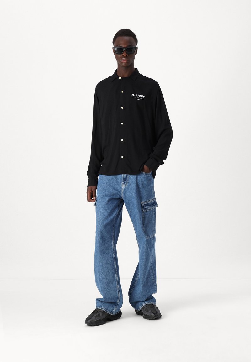 Zwarte lange mouwen knop-up shirt met wit logo, gecombineerd met lichtblauwe cargobroek met zijzakken en zwarte chunky schoenen.