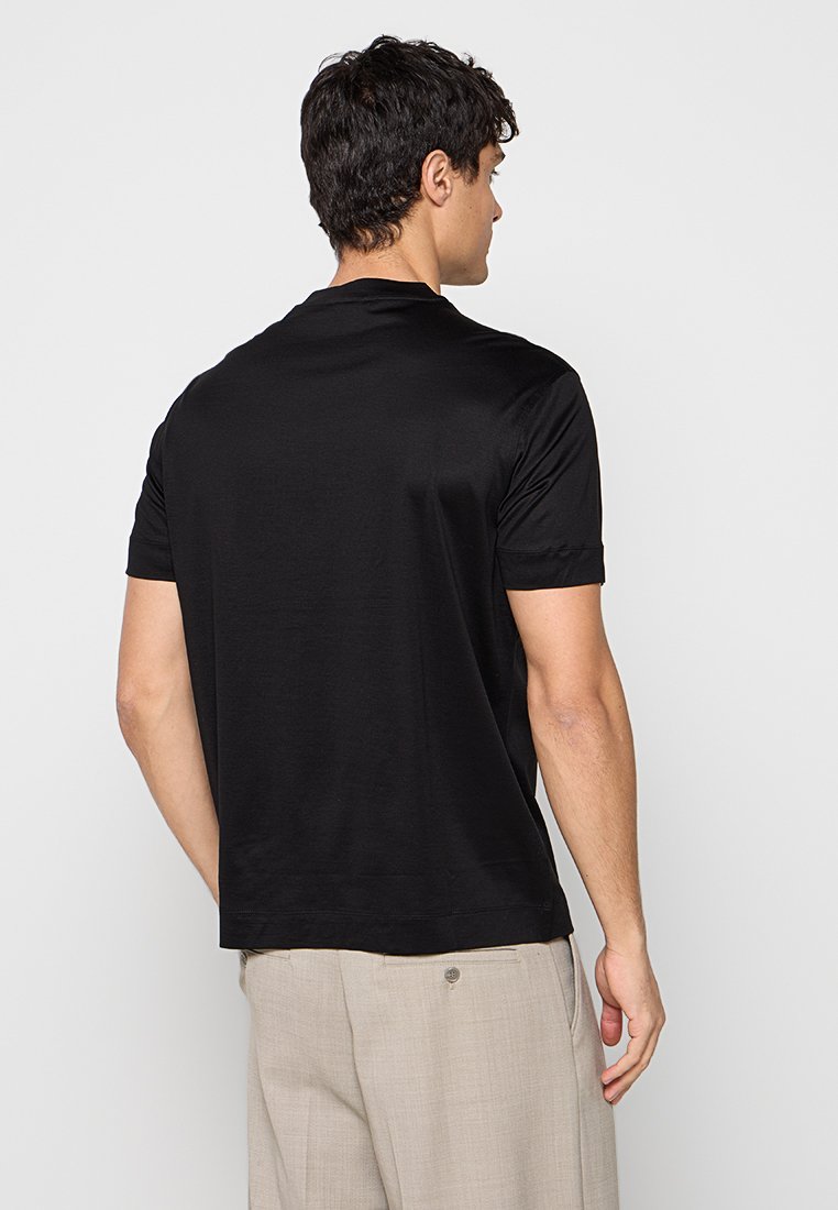 Hombre con cabello oscuro que lleva una camiseta negra de manga corta y pantalones beige, de pie de espaldas sobre un fondo liso.