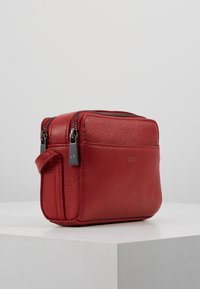 Punainen nahkalaukku, jossa on teksturoitu pinta, kaksinkertaiset vetoketjut yläosassa, sivukahva ja hillitty logo edessä. Kompakti suorakulmainen muotoilu.