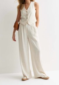 Gilet rayé beige avec un décolleté plongeant en V, quatre boutons, associé à un pantalon large. Le mannequin tient un sac à main marron et porte des baskets neutres.