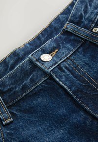 Primer plano de vaqueros vaqueros azules con botón de metal grabado con "NEXT DENIM SINCE 1982" y costura naranja visible en la trabilla y la pretina.