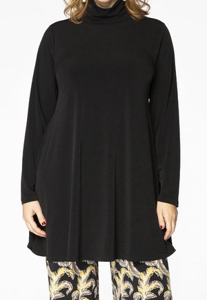 Femme portant une tunique noire à manches longues col roulé et un pantalon noir avec des motifs feuillus jaunes et blancs, debout sur un fond blanc.