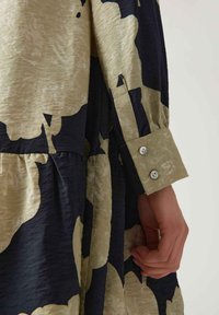 Hand an der Seite ruhend, gekleidet in einem schwarz-beigen blumenmuster langen Kleid mit einem Bund, der zwei Knöpfe hat.