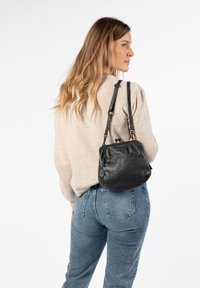 Borsa a tracolla in pelle nera con forma arrotondata, dotata di una tracolla intrecciata, dettagli metallici e un charm con logo. Indossata in modo casual con jeans e un maglione.