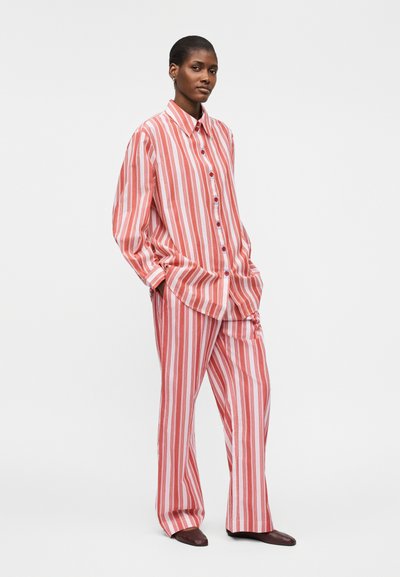 Naine seisab käed taskutes, seljas lahtine punase ja valge vertikaalsete triipudega nööbitav särk ning sobivad laiade pükstega ja pruunide kingadega.