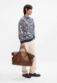 Καφέ δερμάτινη τσάντα duffel με μαλακή υφή, μεγάλη χωρητικότητα και κυλινδρικές λαβές. Συνδυασμένη με πουλόβερ σε σχέδιο μπλε χρώματος και κρεμ παντελόνι.