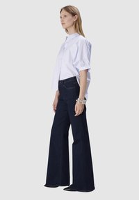 Chemise blanche à boutons avec manches courtes, associée à un jean évasé bleu foncé. Le jean a une texture lisse et un design classique à cinq poches.