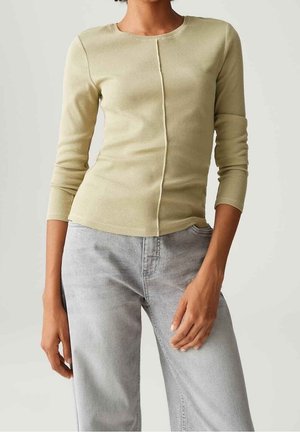 Vrouw draagt een lichtbeige geribd shirt met lange mouwen en een centrale naad, gecombineerd met lichtgrijze hoge taille jeans.