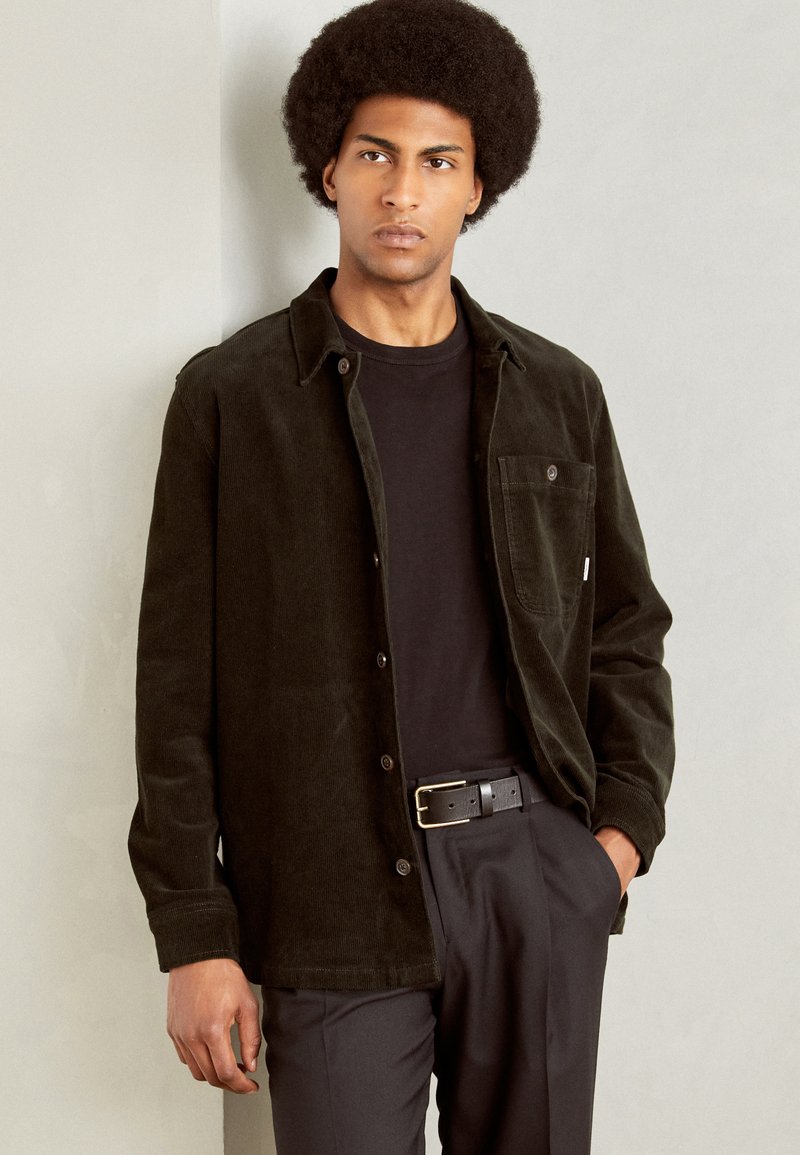 Chaqueta de camisa de pana verde oscuro con cuello de camisa, bolsillo frontal y botones negros, llevada sobre una camiseta negra y pantalones negros.