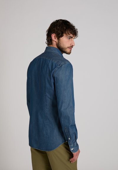 Hombre con cabello rizado oscuro, vestido con una camisa de mezclilla azul y pantalones verde oliva, mirando de lado contra un fondo claro y liso.