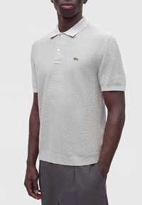 Homme portant un polo à manches courtes gris clair avec un petit logo crocodile vert sur la poitrine gauche et un pantalon gris foncé.